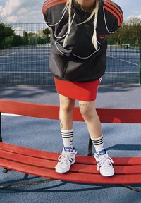 Svart zip-up jacka med röda accenter, röd kjol, vita högsidor sneakers med blå detaljer och randiga strumpor. Sitter på en röd bänk.