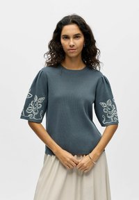 Femme aux cheveux bouclés et foncés portant un haut bleu-gris à manches courtes avec une broderie florale blanche sur les manches et une jupe beige clair, mains jointes.