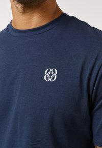 Man wearing a navy blue crew neck t-shirt with a small white embroidered logo on the chest featuring stylized overlapping letters.