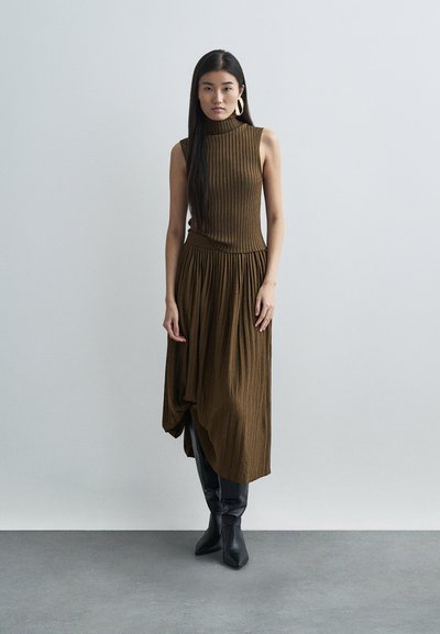Brown ribbed dress with high collar, pleated skirt, and asymmetric hem, paired with knee-high black boots. Smooth texture, fitted design.