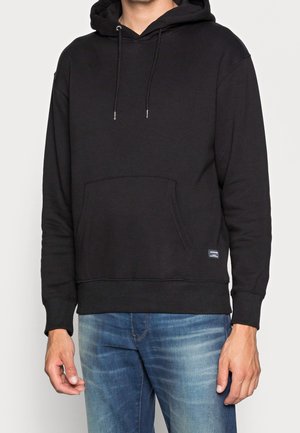 Mand iført ensfarvet sort hættetrøje med forlomme og blå jeans, stående mod en lys baggrund.