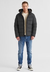 Gepolsterte schwarze Jacke mit Kapuze, featuring einen Reißverschluss und zwei Seitentaschen. Kombiniert mit hellblauen Jeans und schwarzen High-Top-Sneakern.