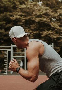 Athlete in a gray tank top and black shorts, wearing a white mesh cap, preparing to sprint on a red track with exercise equipment in the background.