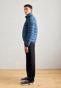 Blauwe pufferjas met doorgestikte textuur, hoge kraag en een merkpatch; gecombineerd met donkere broek en meerkleurige sneakers.