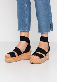 Schwarze Keilsandalen mit Korkplattform und elastischen Riemen, mit offenem Zehendesign und strukturiertem Oberfläch. Jeans mit ausgefranstem Saum.