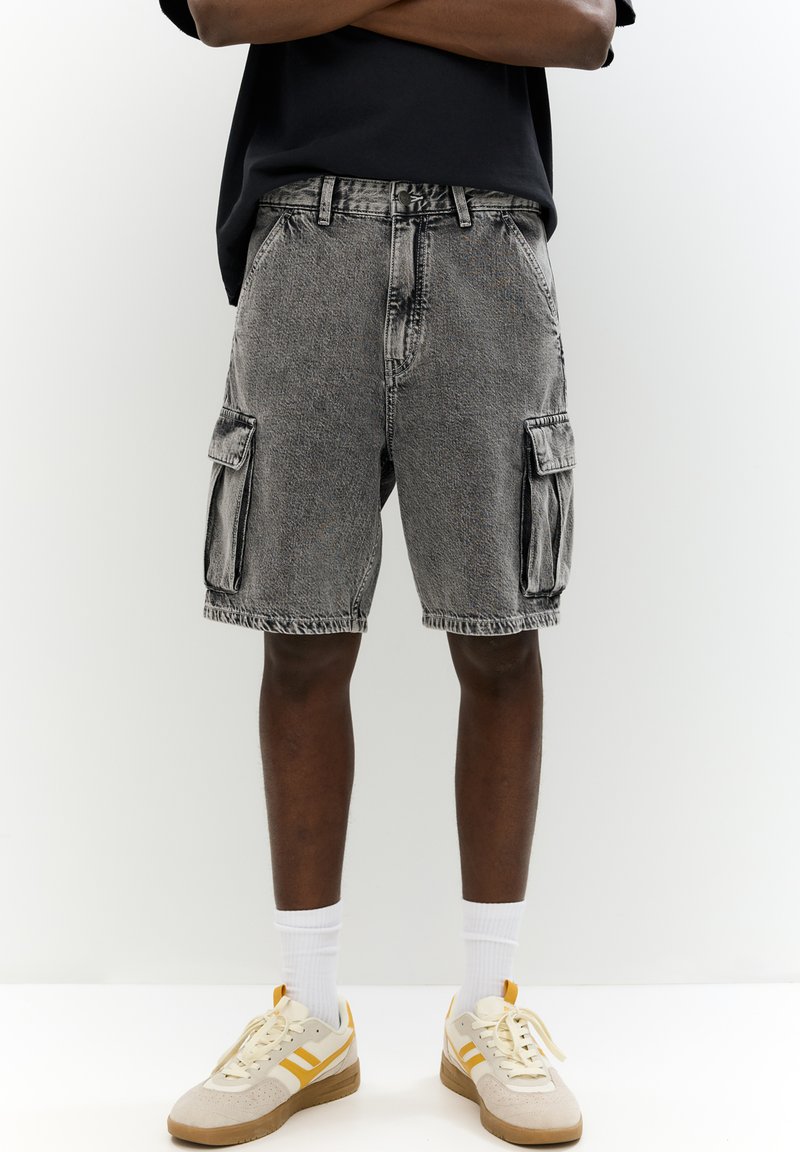 Shorts cargo en denim gris avec des ourlets effilochés, poches latérales et une coupe décontractée, assortis à des chaussettes blanches et des baskets beiges.