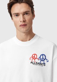 Hombre vestido con una camisa blanca con símbolos de paz rojos y azules, sosteniendo las manos sobre el texto "ALLSAINTS" y "MENSAJE A UN FUTURO".
