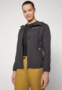 Svart utendørsjakke med glidelås i front, høy krage og meshinspirerte detaljer. Kombinert med gule bukser som har snøring og en slim fit.
