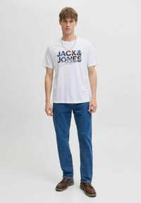 Weißes T-Shirt mit kurzen Ärmeln und einem bunten Drucklogo mit der Aufschrift "JACK & JONES" auf der Vorderseite, kombiniert mit blauen Jeans und braunen Schuhen.