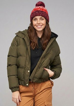 Smiling woman with long brown hair wearing a red knit hat, olive green puffer jacket, black hoodie, and tan cargo pants against a plain background.