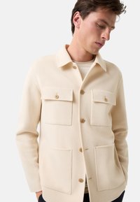 Beżowa koszula z wełny z dodatkiem innych włókien, z kołnierzykiem, dwiema kieszeniami na piersi oraz trzema guzikami z przodu. Gładka faktura z minimalistycznym designem.