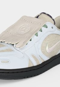 Λευκό και μπεζ αθλητικό παπούτσι Nike με πράσινα κορδόνια, μαύρη σόλα, ιριδίζον Swoosh και δερμάτινη φτερούγα με κρόσσια πάνω από την κορυφή.