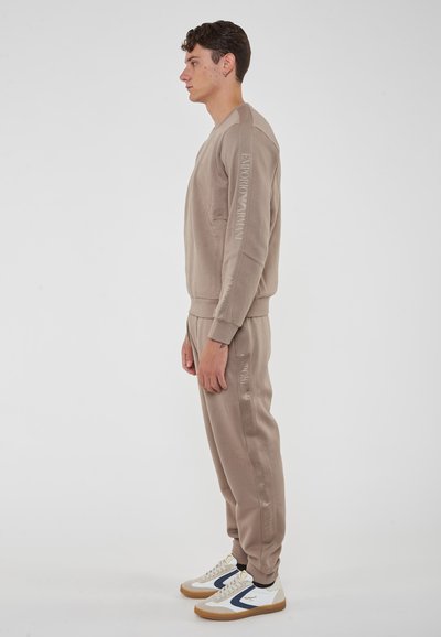 Completo di felpa e pantaloni beige con polsini a coste. Logo "Emporio Armani" stampato verticalmente sulla manica e sulla gamba. Sneakers con suola in gomma.