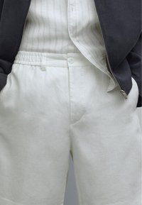Shorts en lin blancs avec poches latérales et fermeture à bouton, associés à une chemise à rayures claires et une veste gris foncé.