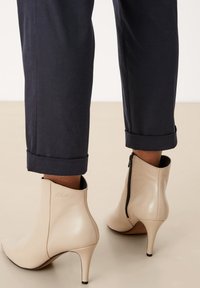 Béžové kotníkové boty s špičatou špičkou a jehlovým podpatkem. Lesklý kožený materiál se bočním zipem a jemným detailem loga.