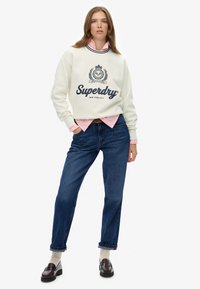 Sweatshirt crème avec logo en texte marine, superposé à une chemise rose clair. Jean en denim bleu avec les poignets retroussés, associé à des mocassins foncés et des chaussettes beige.