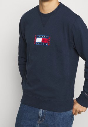 Mężczyzna ubrany w granatową bluzę z naszywką z logo Tommy Jeans na piersi oraz niebieskie dżinsy, z ręką w kieszeni.
