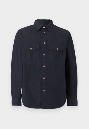 Fiatal férfi áll, fekete gombos inget, világoskék bő szárú farmert és fehér tornacipőt visel, egyszínű világos háttér előtt.