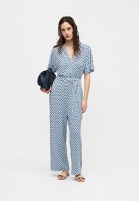 Γυναίκα που φοράει μπλε ολόσωμη φόρμα με σχέδια, κρατώντας μια μπλε υφή τσάντα χειρός, με μπεζ σανδάλια ανοιχτής μύτης, στέκεται μπροστά σε μονόχρωμο φόντο.