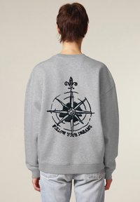 Grijze sweatshirt met een zwart geborduurd kompasontwerp en de tekst "VOLG JE DROMEN" op de achterkant. Heeft een relaxte pasvorm en geribde manchetten.