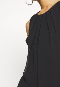 Robe noire sans manches mettant en avant de douces plis, un tissu fluide et des accents transparents sur les côtés, créant une silhouette ample et élégante.