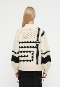 Striktrøje med en cremet bundfarve og sorte geometriske mønstre, ribbet kant og ballonsleeves. Tekstur detajler forbedrer designet.