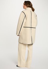 Abrigo crema con líneas geométricas negras, tela texturizada, cuello alto; combinado con pantalones de pana beige. Modelo visto desde la espalda.