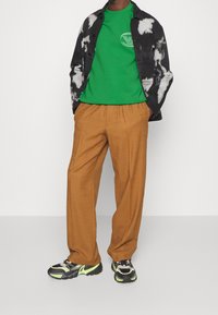 Maglietta tie-dye bianca e nera sopra una t-shirt verde con logo, pantaloni beige rilassati e sneakers nere con dettagli neon.