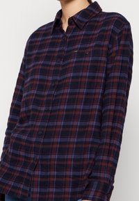 Camicia in flanella a quadri nei colori blu navy, borgogna e blu. Presenta una chiusura con bottoni sul davanti, colletto classico e due tasche sul petto. Texture morbida.
