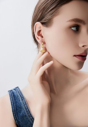 Gouden oorringen met een waaiervormig hangerdetail en een kleine kristalaccent, gedragen door een model met een denim outfit.