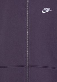 Ljubičasta zip-up dukserica izrađena od mekanog materijala, sa prednjim zatvaračem i bijelim Nike logotipom izvezenim na lijevoj strani grudi.