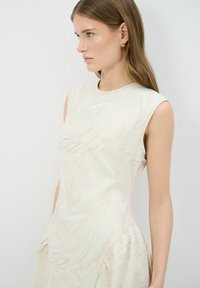 Crème jurk met een gestructureerd, golvend patroon. Mouwloos ontwerp en aansluitende silhouet, met subtiele zijden plooien voor extra detail.