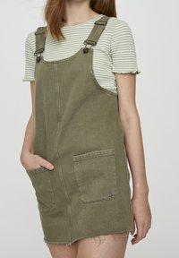 Robe salopette en denim vert olive avec bretelles réglables, deux poches avant et ourlet effiloché. Superposée sur un t-shirt rayé vert clair.
