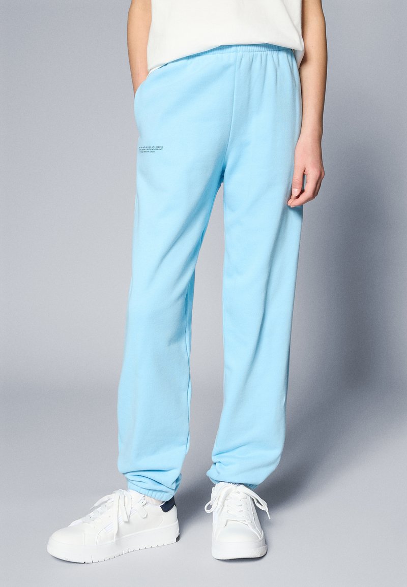Pantalones de chándal de algodón suave en azul claro, con cintura elástica y bolsillos laterales. Presentan texto impreso en la pierna izquierda. Combinados con zapatillas blancas.