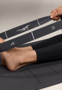 Čierny elastický odporový pás držený medzi nohami na tmavej textúrovanej jogovej podložke. Pás má číslované sekcie a šedý dizajn postavy.