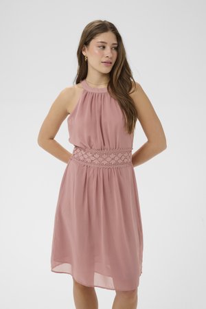 Jonge vrouw in een mouwloze stoffige roze jurk met kanten tailleband, handen achter de rug, kijkt naar rechts tegen een effen lichte achtergrond.