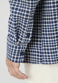 Manche de chemise à carreaux bleus et blancs avec poignet boutonné, présentant un tissu texturé et une coupe ajustée.