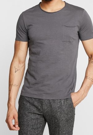 Mand iført en tætsiddende mørkegrå t-shirt med korte ærmer og brystlomme samt teksturerede grå bukser, med tatoveringer på begge underarme.