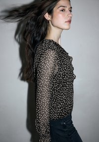 Zwarte doorschijnende blouse met wit bloemmotief, gerafelde voorkant en lange mouwen. Gecombineerd met donkere spijkerbroek. Gladde textuur.