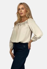 Beżowa bluzka z haftowanym koronkowym wykończeniem przy dekolcie i mankietach, z bufiastymi długimi rękawami, zestawiona z ciemnymi dżinsami. Gładki materiał, luźny krój.
