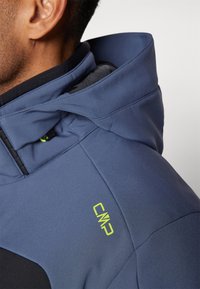 Chaqueta softshell grisazul con cuello alto, cremallera visible y logo "CMP" en neón verde en el hombro. Tejido texturizado y diseño ajustado.