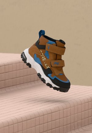 Brown, blue, and black hiking shoe with GORE-TEX label and Velcro straps, hovering above pink tiled stairs against beige background.