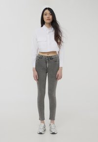Camicia bianca cropped con bottoni e due tasche sul petto, abbinata a jeans skinny grigi e sneaker atletiche argentate. Tessuto liscio e struttura ben definita.