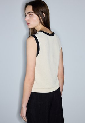 Giovane donna con lunghi capelli castani che indossa una canotta color crema con rifiniture nere e pantaloni neri larghi, rivolta verso sinistra su uno sfondo semplice.