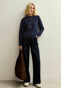 Maglione navy, pantaloni in denim scuri a gamba larga, sneakers crema e borsa a spalla in suede marrone; texture dei tessuti lisci e elementi di design semplici.