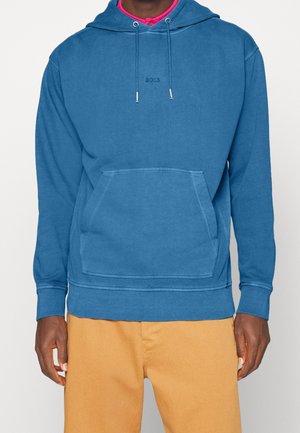 Mandlig model iført en blå BOSS hættetrøje med en forlomme og beige bukser, stående foran en ensartet lys baggrund.