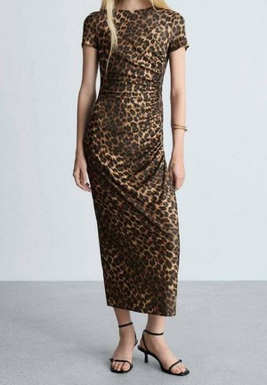 Frau trägt ein figurbetontes, kurzärmeliges, knöchellanges Kleid mit Leopardenmuster und schwarze, Riemchen-Heels, steht vor einem schlichten Hintergrund.