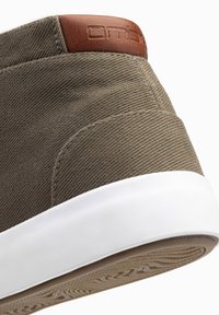 Zapatillas de lona verde oliva con suela blanca, que presentan un acento de cuero marrón en el talón y costuras texturizadas para un detalle adicional.