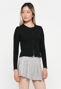 Svart ribbet cardigan med blondekant langs halsen og knappelukking. Kombinert med et lys grått plissert skjørt.