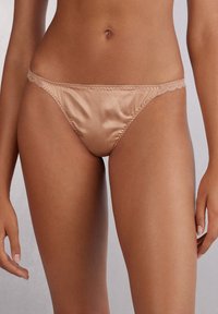 Satinstringtrosor i en varm beige nyans med delikat spetsdetalj längs midjan, med en mjuk yta och minimal täckning.